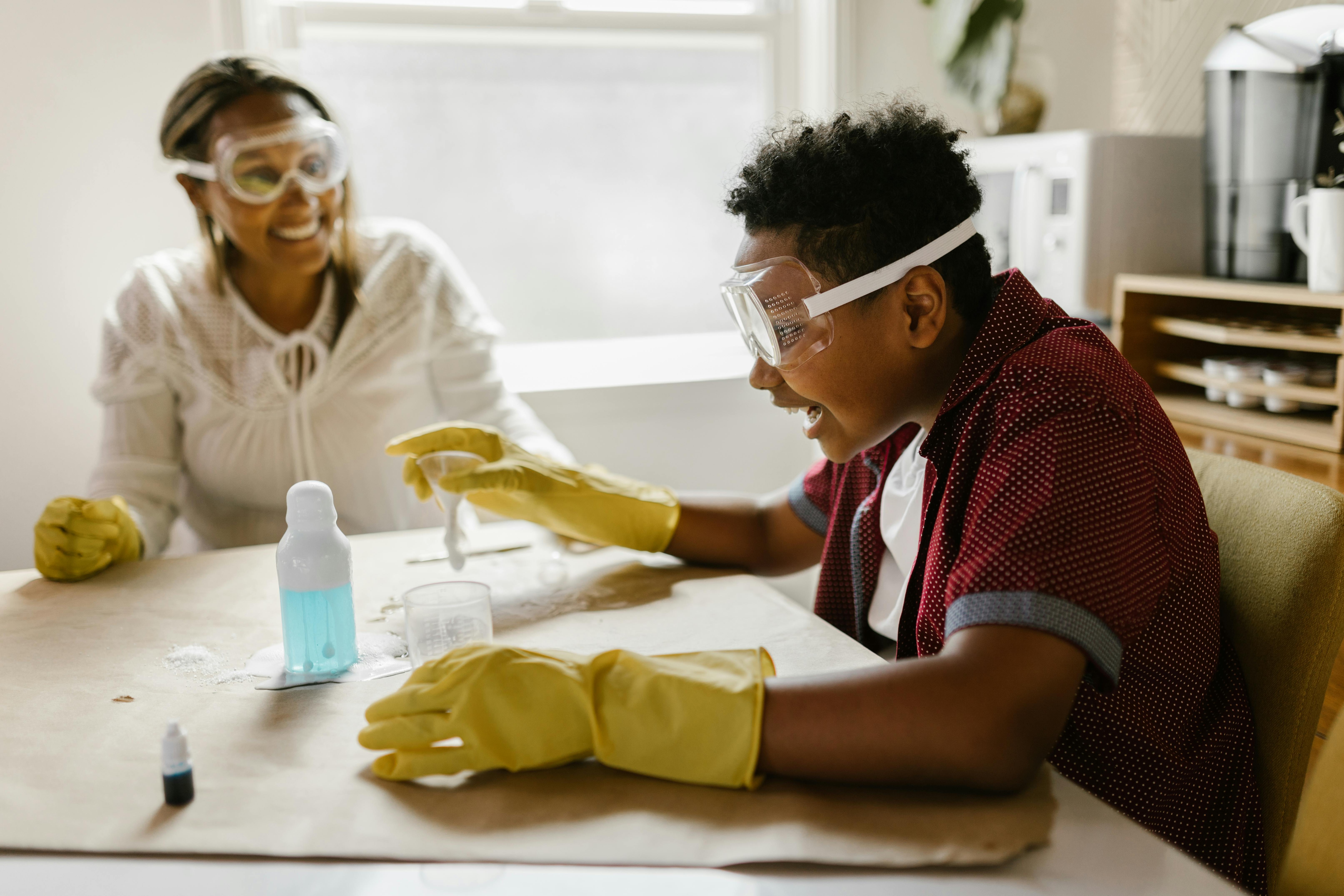 Enfant qui sourit en faisant une expérience scientifique, on aperçoit sa mère qui rit en arrière plan. Photo RDNE Stock project