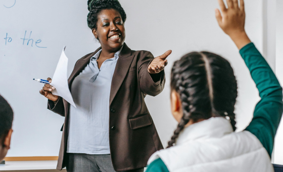 Une enseignante, au tableau, interroge en souriant une élève que l'on voit de dos.