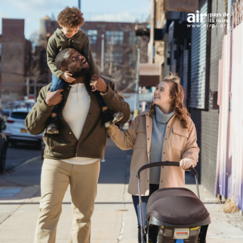 Trois personnes se promenant en ville, elles sourient. L'une porte un enfant sur ses épaules et l'autre pousse une poussette